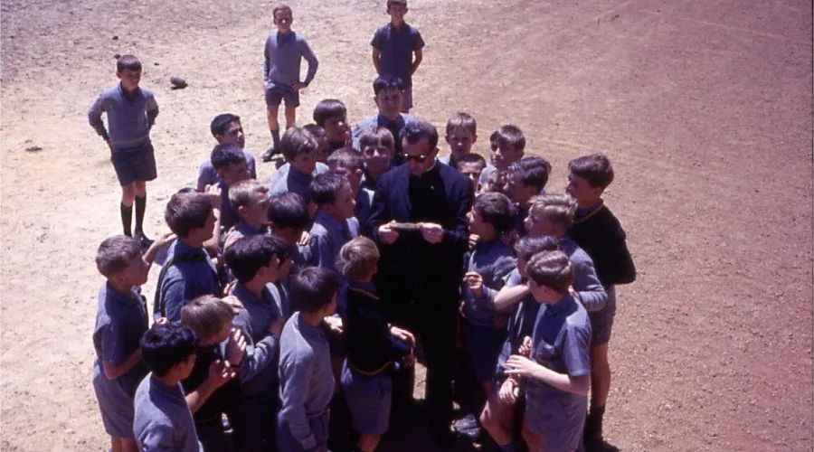 A heritage photo of students gathering around a priest to listen to an announcement at Mazenod College Perth WA
